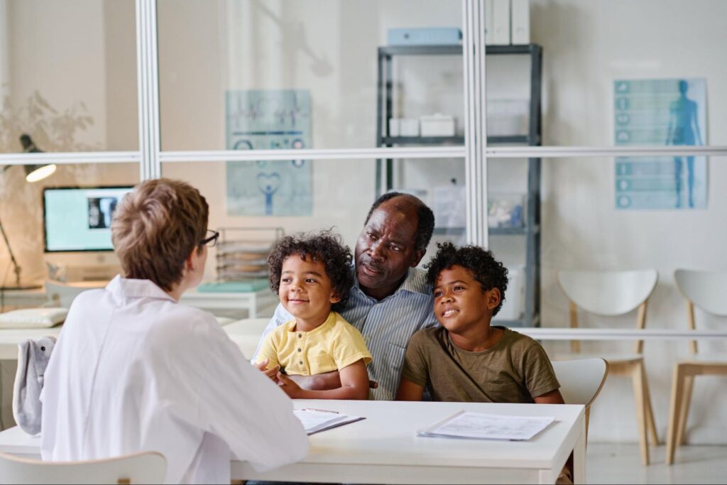 Man sitting in a doctor's office with two children and a woman in a white coat sitting across from them.